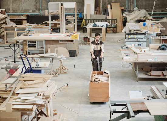 Young worker of modern factory pushing cart with large box full of leftovers of workpieces while moving along workshop